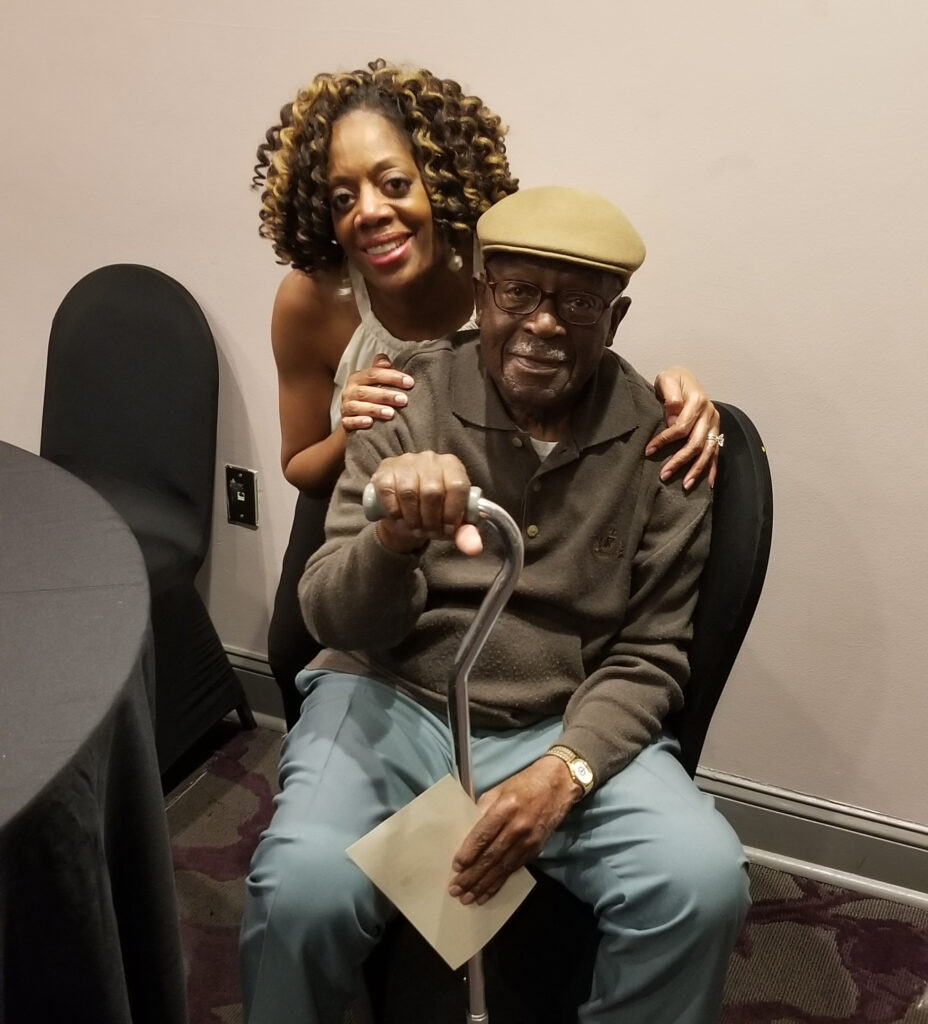 A black woman hugging an elderly black man wearing a tan cap, brown shirt and blue pants.